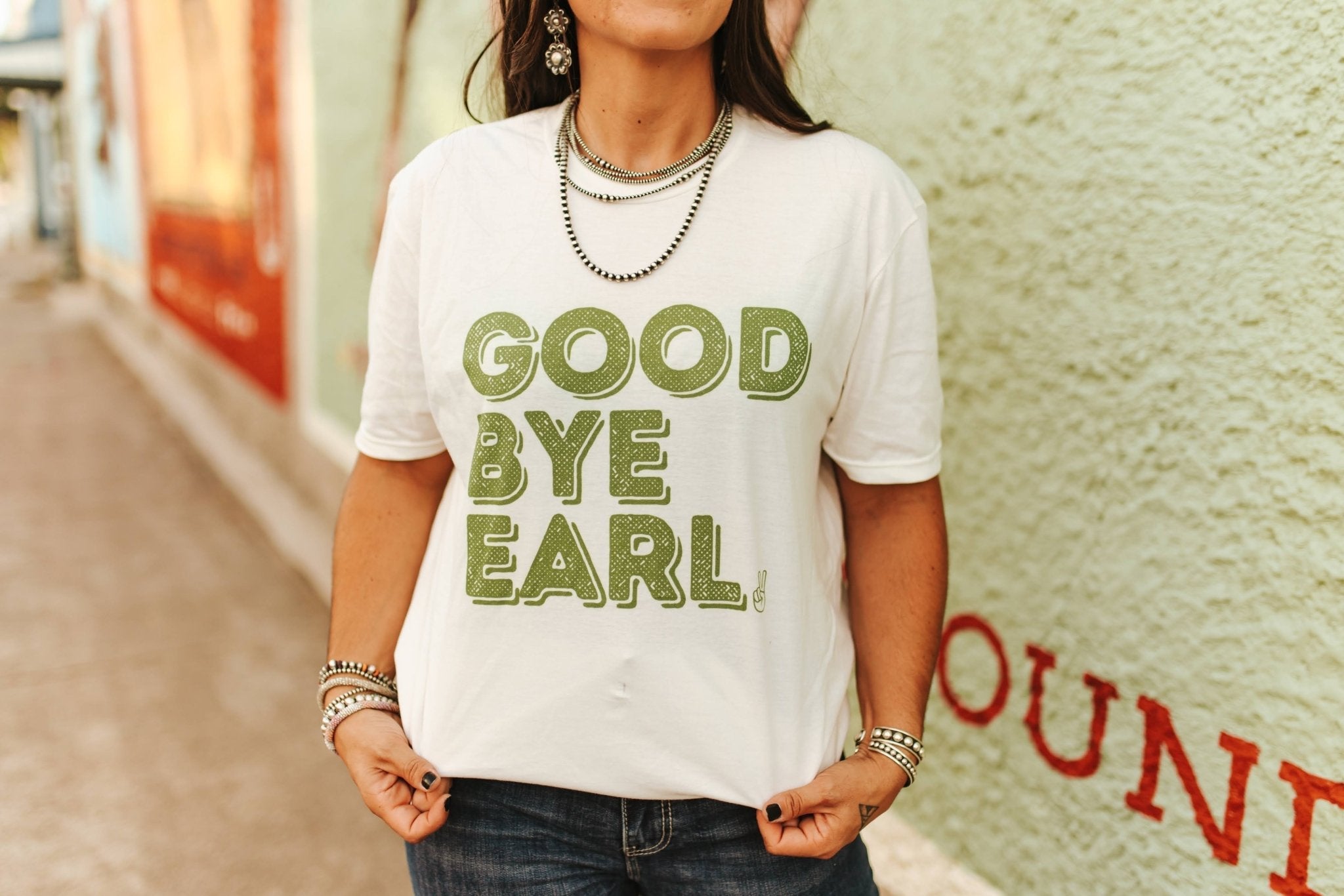 Person wearing a white t-shirt with 'GOOD BYE EARL' text, standing against a textured wall.