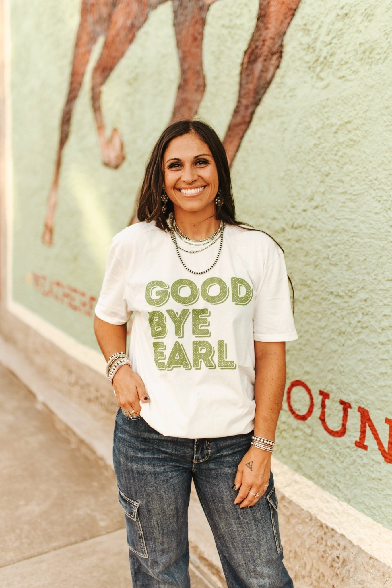 Woman wearing a 'Good Bye Earl' t-shirt standing in front of a mural.