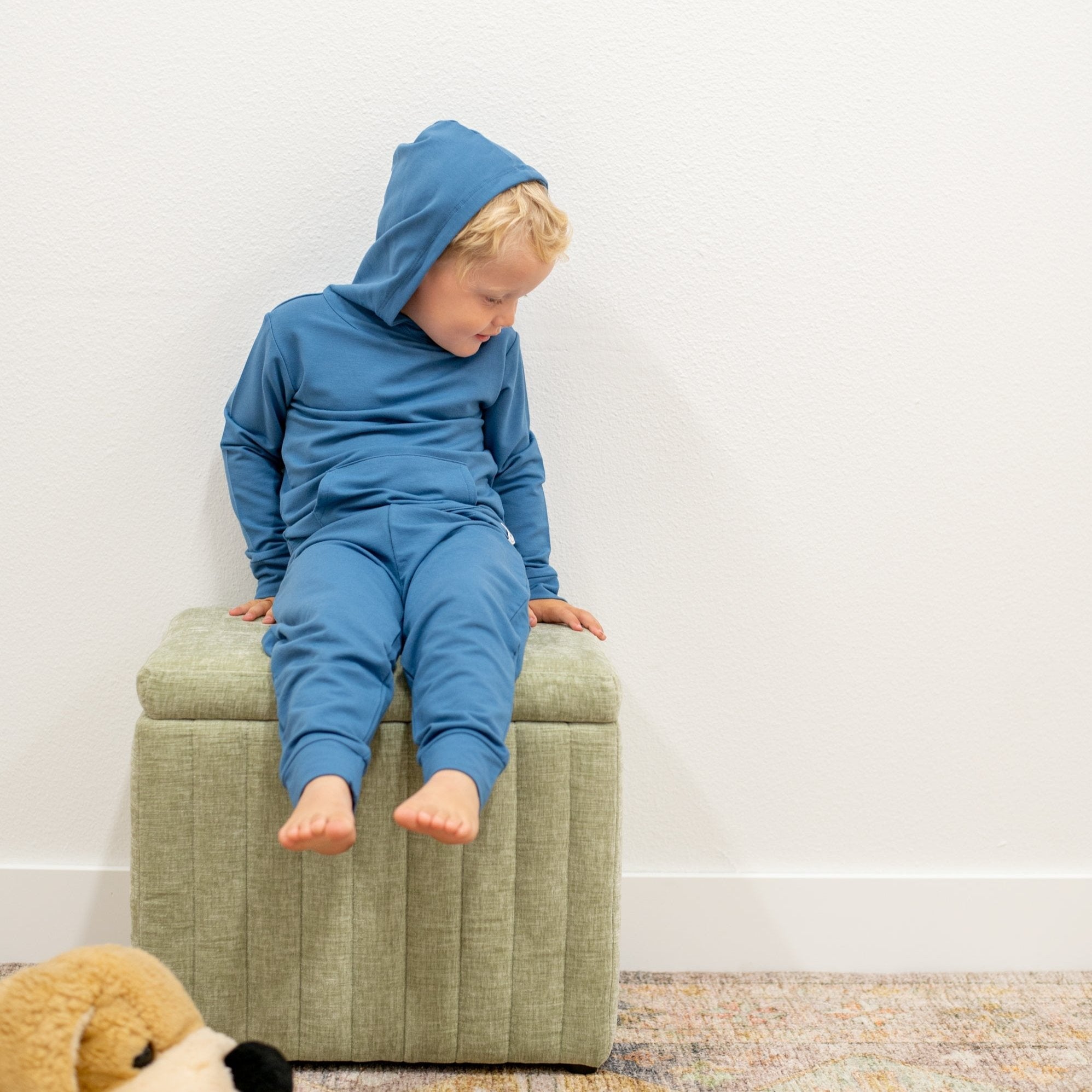 Child wearing a blue hoodie and pants sitting on a green ottoman against a white wall.