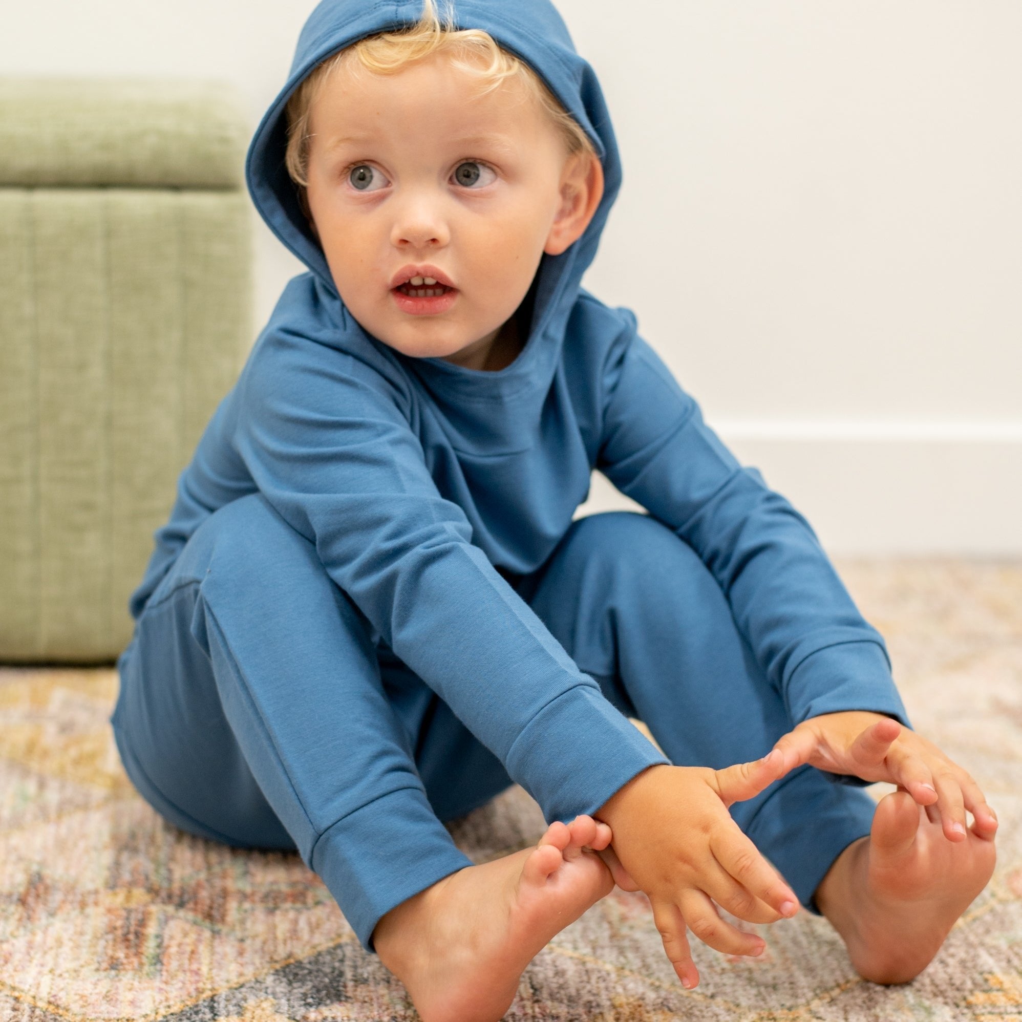 Child wearing a blue hoodie and pants sitting on a carpeted floor.