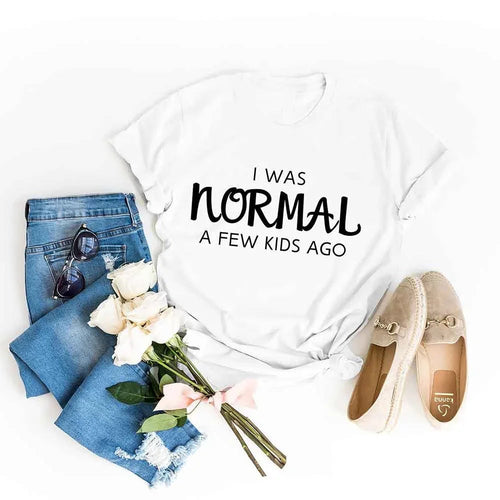 White t-shirt with 'I was normal a few kids ago' text, blue jeans, sunglasses, flowers, and shoes on a white background