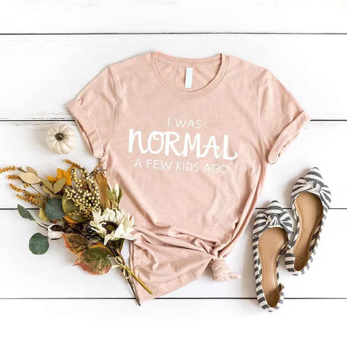 Pink t-shirt with 'I was normal a few kids ago' text, surrounded by flowers and shoes on a wooden surface.