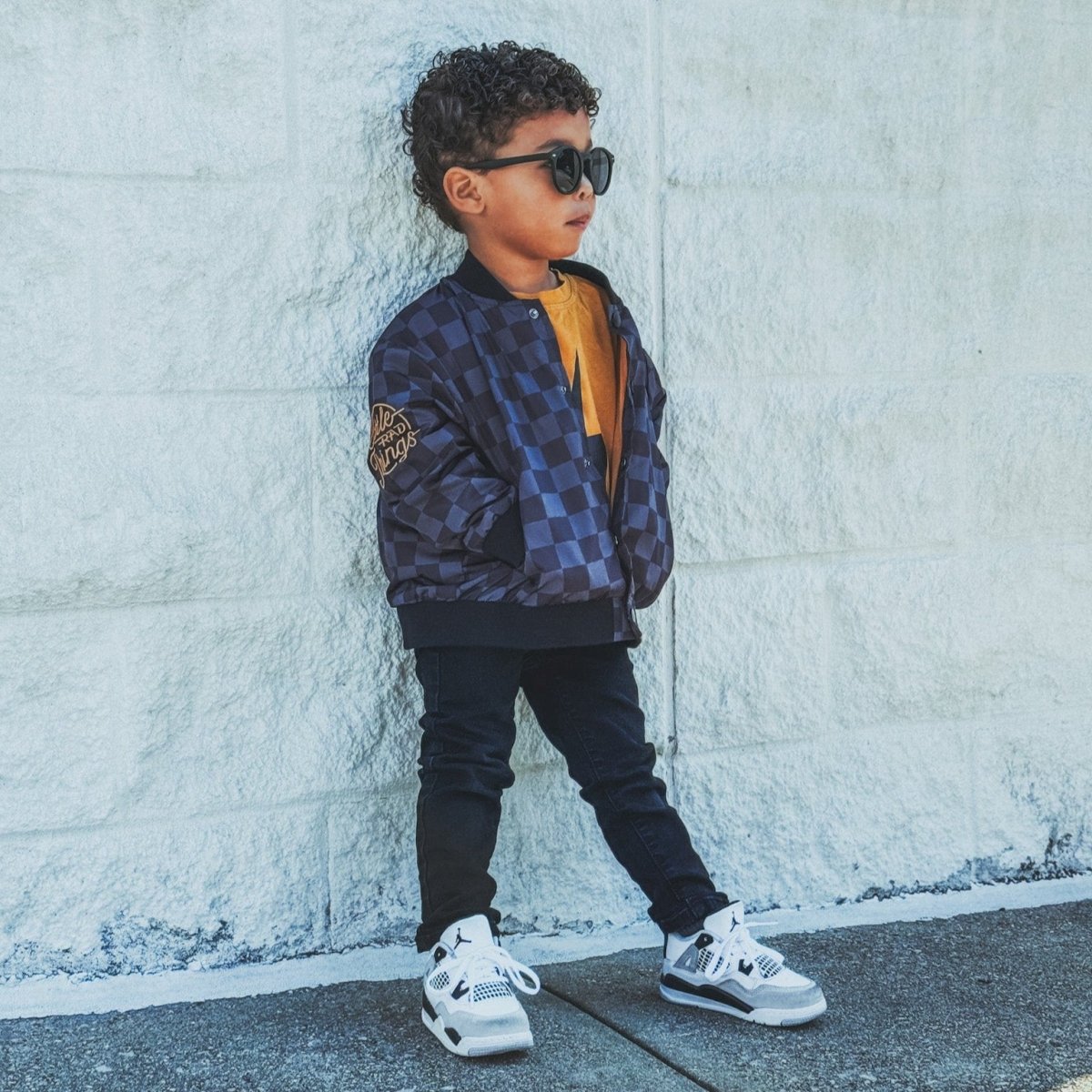 Child wearing a blue checkered jacket and sunglasses leaning against a white wall.