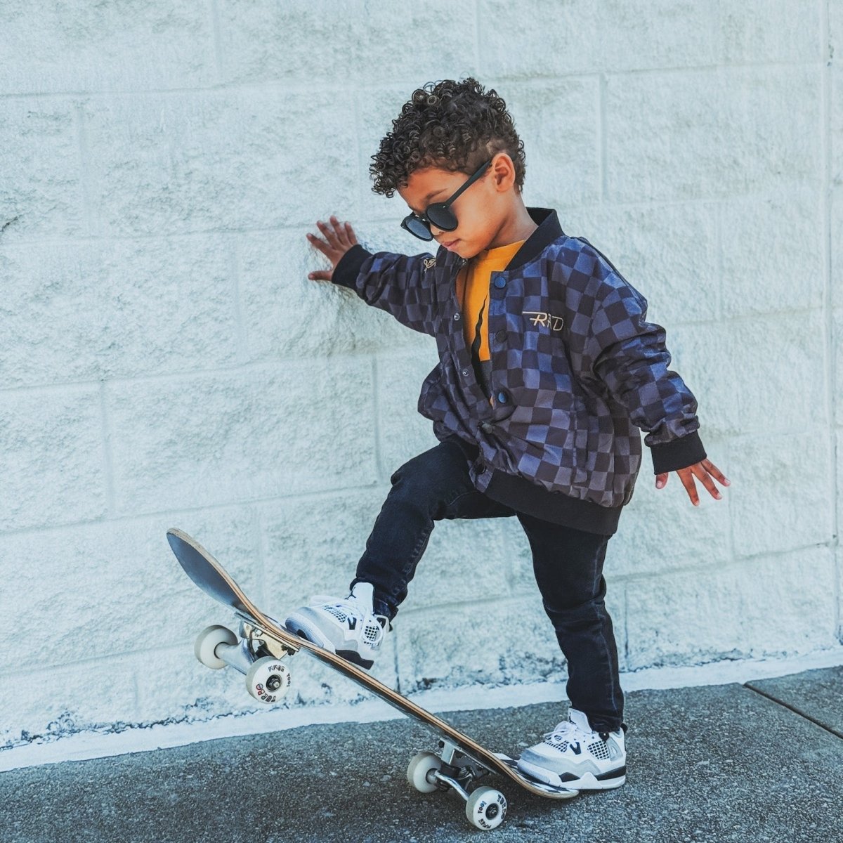 Child in a checkered jacket and sunglasses standing on a skateboard against a light gray wall.