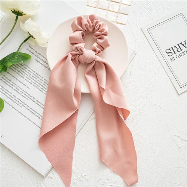 Pink satin hair scrunchie with a bow on a white surface with flowers and text.