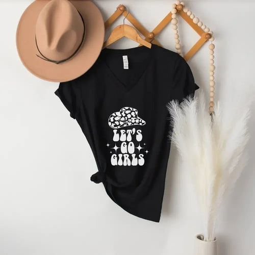A black V-neck T-shirt hanging on a wooden hanger against a white wall, featuring a white graphic of a flower crown and the text 'LET'S GO GIRLS'. A tan wide-brim hat is draped over the left shoulder, and decorative wood beads and pampas grass hang on the right.