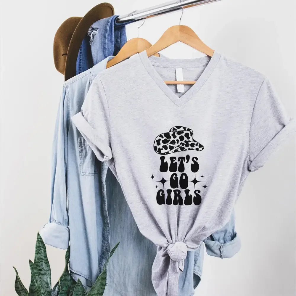Gray t-shirt with 'Let's Go Girls' print on a hanger with denim jacket and hat in the background.