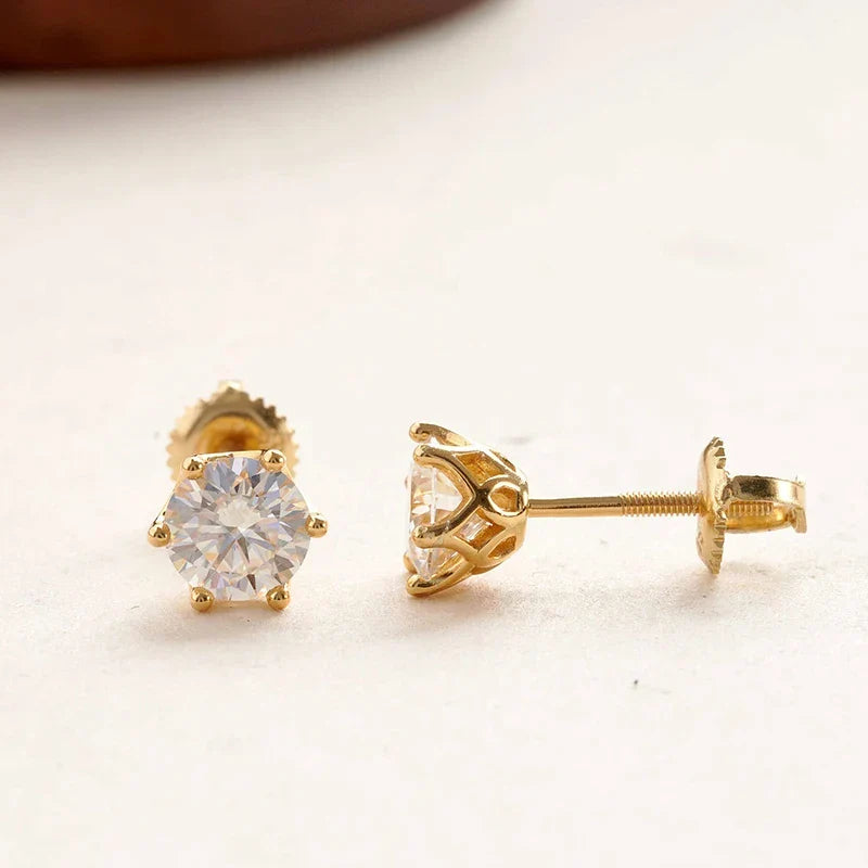 A pair of gold stud earrings, one shown from the front with a round clear gemstone and the other from the side showing the post and butterfly backing, on a light beige background.