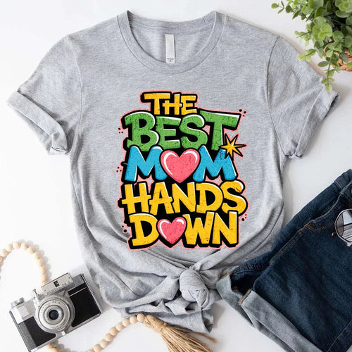 Gray t-shirt with colorful text 'The Best Mom Hands Down' on a white surface with accessories.