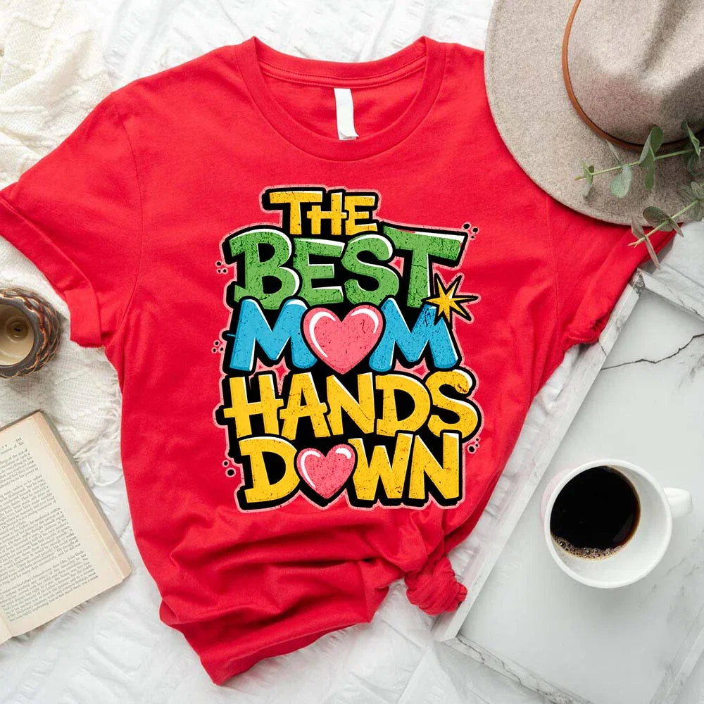 Red t-shirt with colorful text 'The Best Mom Hands Down' on a white surface with a hat, book, and coffee cup.