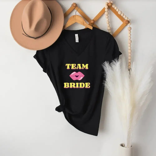 Black t-shirt with 'Team Bride' text and pink lips graphic on a white wall with a hat and decorative elements.
